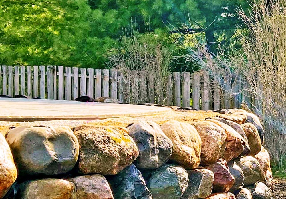Stone retaining wall with wooden fence and lush green trees in the background.