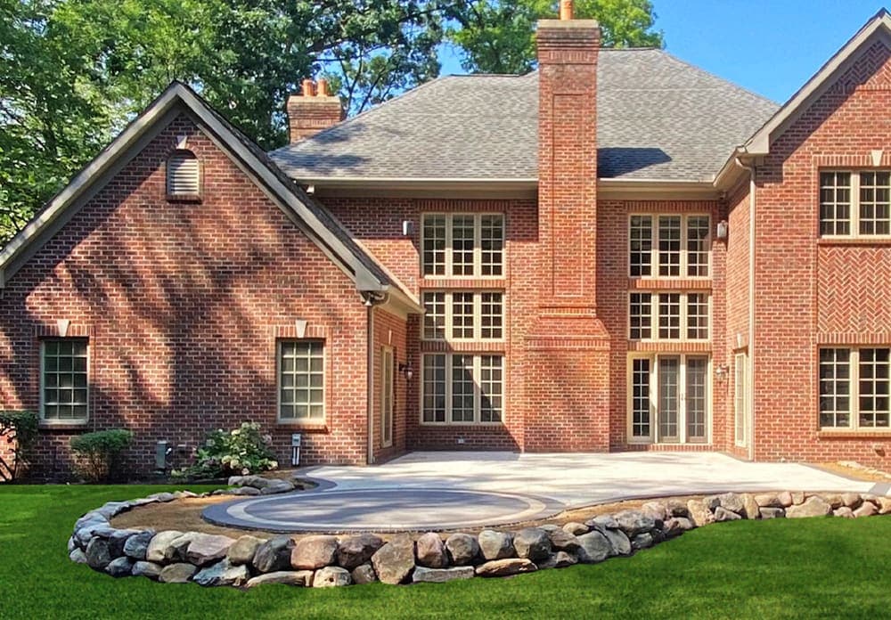 Elegant brick home with landscaped yard and stone patio, surrounded by greenery.