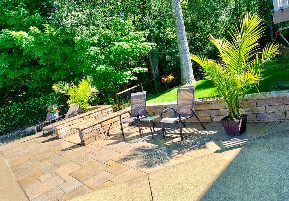 Patio area with chairs, plants, and stone wall, surrounded by lush greenery and sunlight.