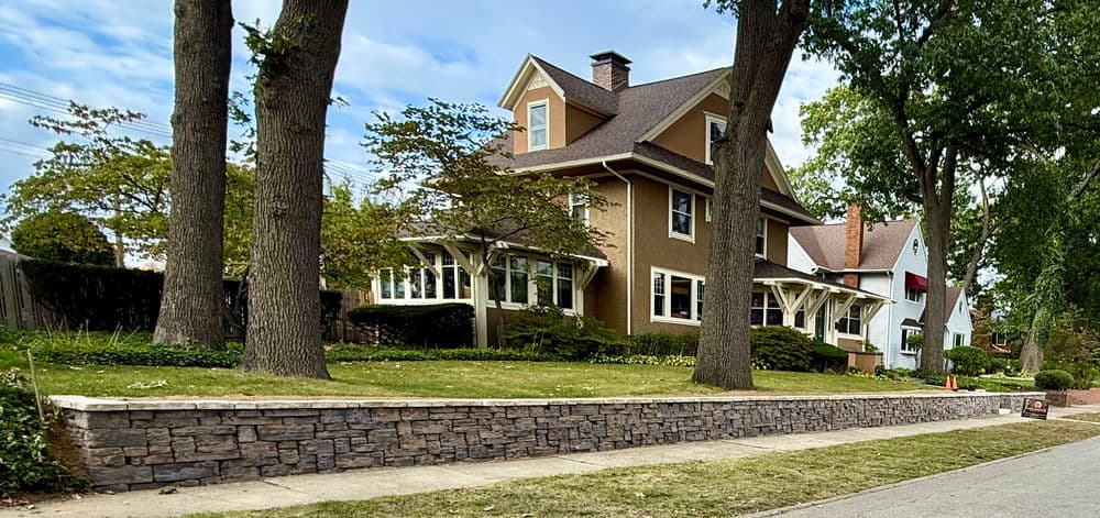 Historic two-story house with garden, surrounded by trees and a stone wall.