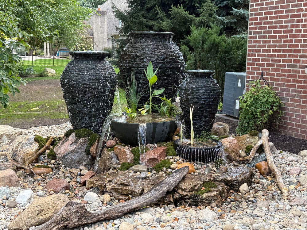 Decorative outdoor fountain with stacked stone pots surrounded by plants and rocks.