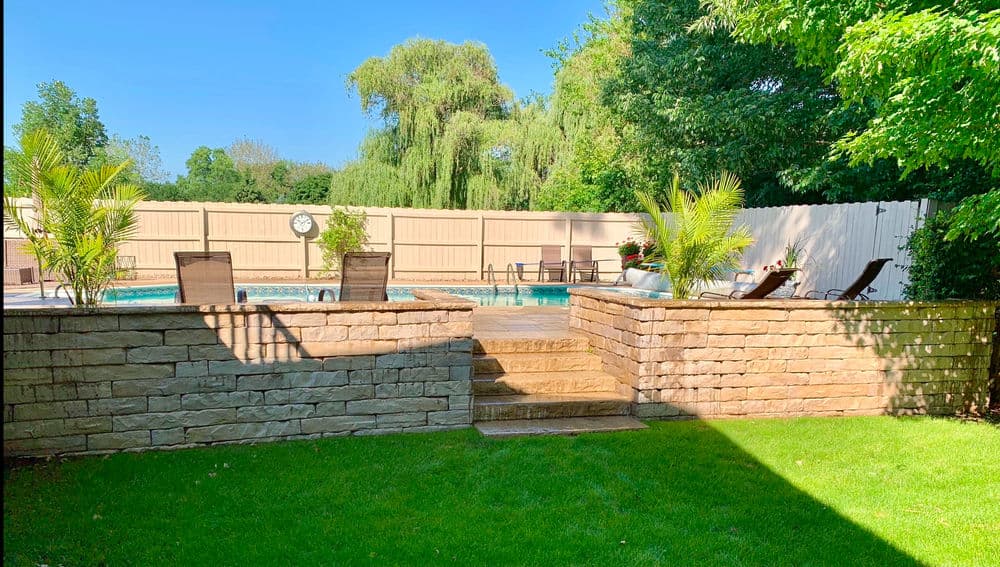 Backyard with stone retaining wall, pool, lounge chairs, and lush greenery under clear blue sky.